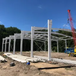 Montaż konstrukcji stalowej hali z użyciem czerwonego dźwigu, widoczna platforma robocza i przygotowane elementy, błękitne niebo w tle.