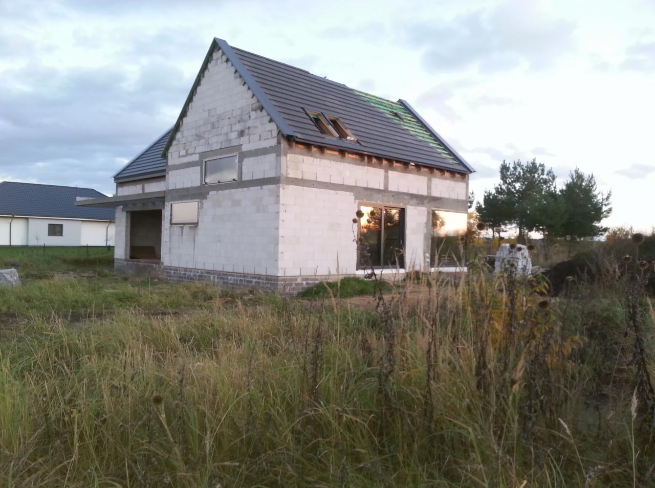 Surowy stan domu jednorodzinnego z białych bloczków, z zamontowanymi oknami i częściowo pokrytym dachem z dachówki ceramicznej, widok z zewnątrz na tle wysokiej trawy i zachmurzonego nieba.