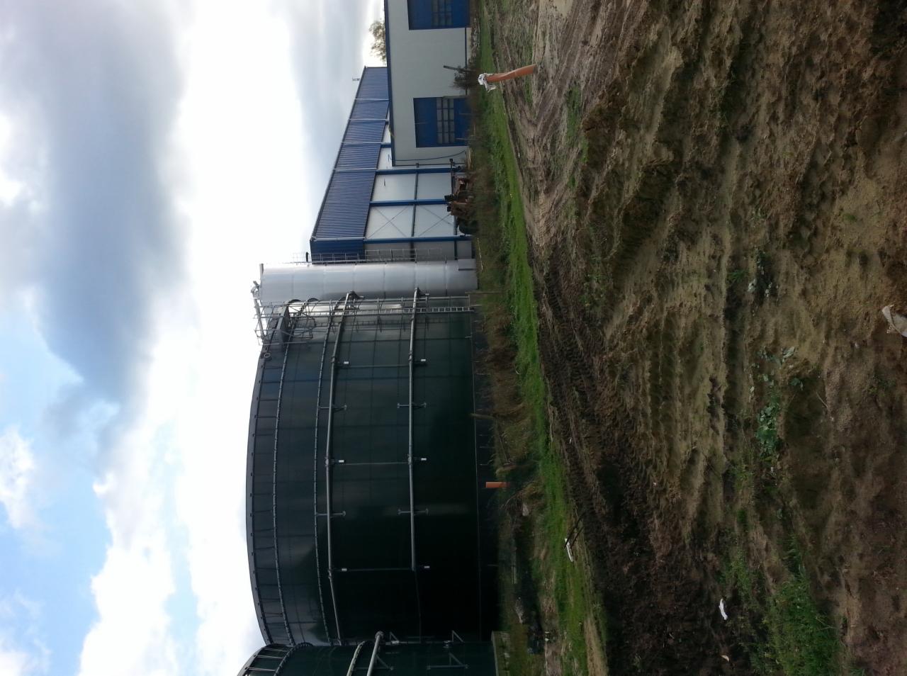 Duży, ciemnozielony zbiornik przemysłowy z zewnętrzną konstrukcją schodów, sąsiadujący z budynkiem magazynowym o niebieskim dachu, na tle pochmurnego nieba i ziemi z widocznymi śladami opon.