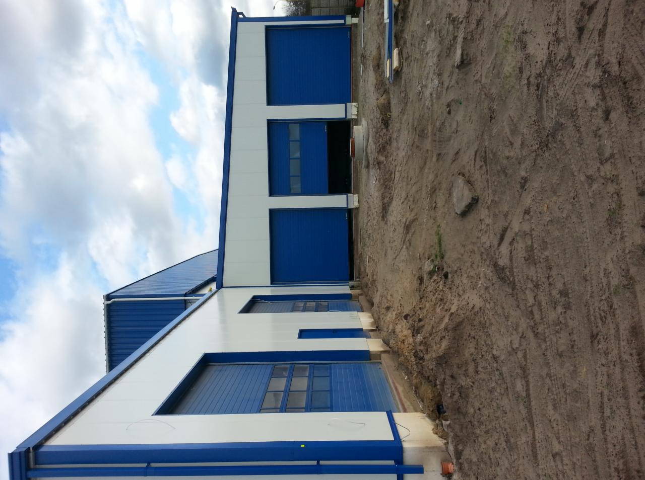 Gotowa hala przemysłowa z niebieskimi bramami segmentowymi i białymi panelami ściennymi, widoczna z zewnątrz na tle pochmurnego nieba i ziemi.