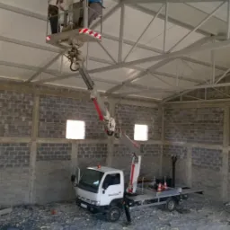 Dw&oacute;ch pracownik&oacute;w na podnośniku koszowym instaluje białe panele pod sufitem wewnątrz hali o konstrukcji stalowej i ścianach z betonowych blok&oacute;w.