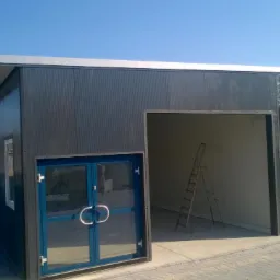 Nowoczesna hala z płyt warstwowych w kolorze grafitowym, z niebieskimi drzwiami wejściowymi i otwartą bramą garażową, drabina wewnątrz, widok z zewnątrz w słoneczny dzień.