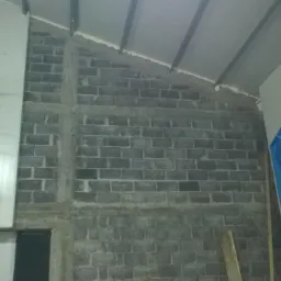 Wnętrze hali w trakcie budowy: murowana ściana z szarych bloczków, częściowo obłożona płytami warstwowymi, widoczne rusztowanie i zabezpieczona niebieską folią płyta.