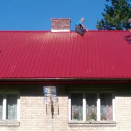 Dwóch dekarzy pracuje na czerwonym, metalowym dachu domu z cegły, widoczny komin i antena telewizyjna.
