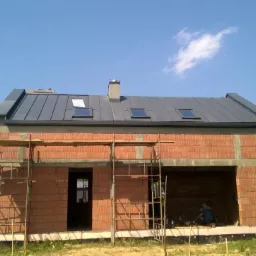 Surowy budynek w trakcie budowy z cegły, z zamontowanym grafitowym dachem z oknami dachowymi, widoczne rusztowania i otoczenie z zielenią.