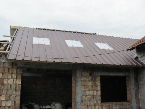 Brązowy dach z blachy trapezowej z trzema świetlikami, częściowo zamontowany na murowanym budynku w trakcie budowy, widoczne elementy więźby dachowej.