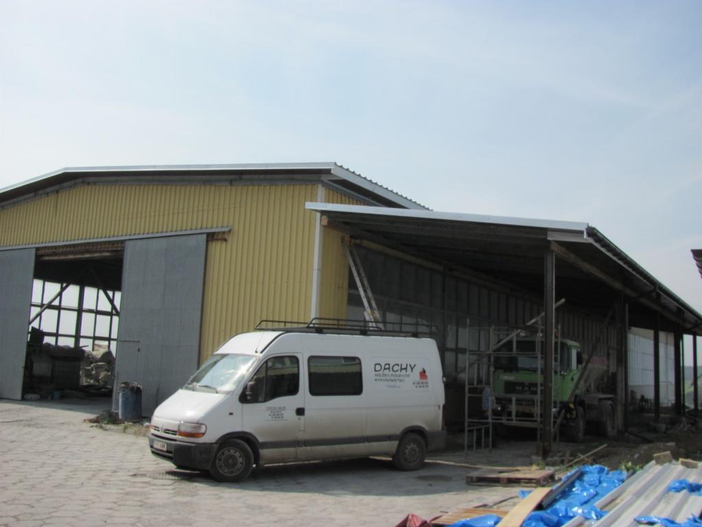 Żółta hala z przesuwnymi drzwiami, biały bus z napisem 'DACHY' zaparkowany przed budynkiem, w tle zadaszona konstrukcja z widocznym sprzętem budowlanym i niebieskimi plandekami.