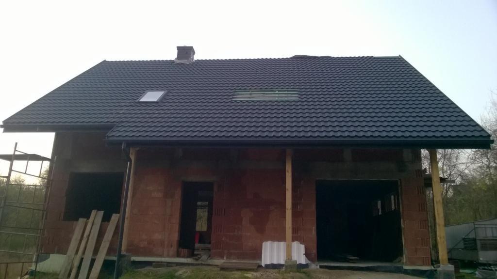 Dom w trakcie budowy z ciemną dachówką, widoczne okna dachowe i komin, ściany z czerwonej cegły, rusztowanie z lewej strony.