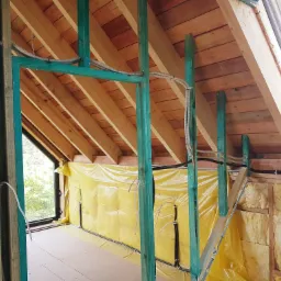 Wnętrze poddasza w trakcie adaptacji, widoczne drewniane elementy konstrukcyjne pomalowane na zielono, izolacja z wełny mineralnej, folia paroizolacyjna w kolorze żółtym oraz poprowadzone kable...