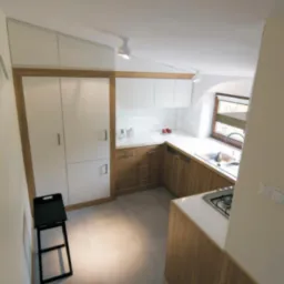 Nowoczesna kuchnia o powierzchni 10 m2 z białymi szafkami górnymi i drewnianymi dolnymi, jasnymi płytkami podłogowymi i czarnym składanym krzesłem.