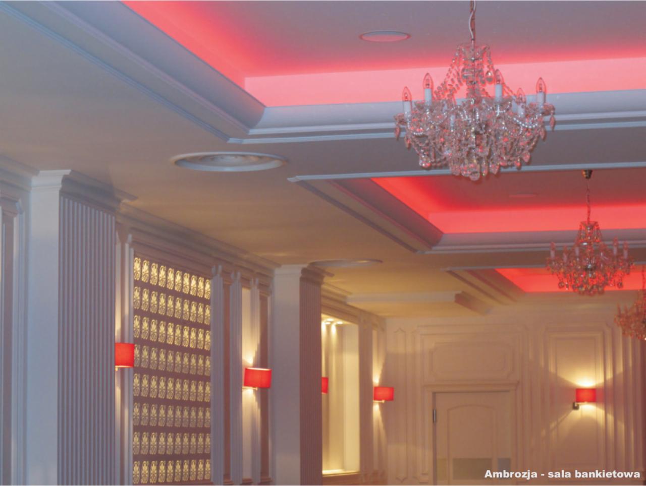 Elegancka sala bankietowa z białymi ścianami zdobionymi sztukaterią, kryształowymi żyrandolami i czerwonym oświetleniem sufitowym.