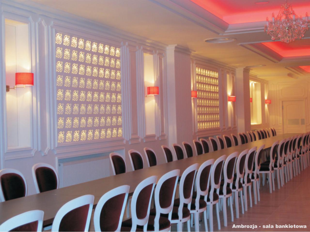 Sala bankietowa z długim stołem i krzesłami, dekoracyjnymi panelami ściennymi z podświetleniem oraz czerwonym oświetleniem sufitowym i żyrandolem.