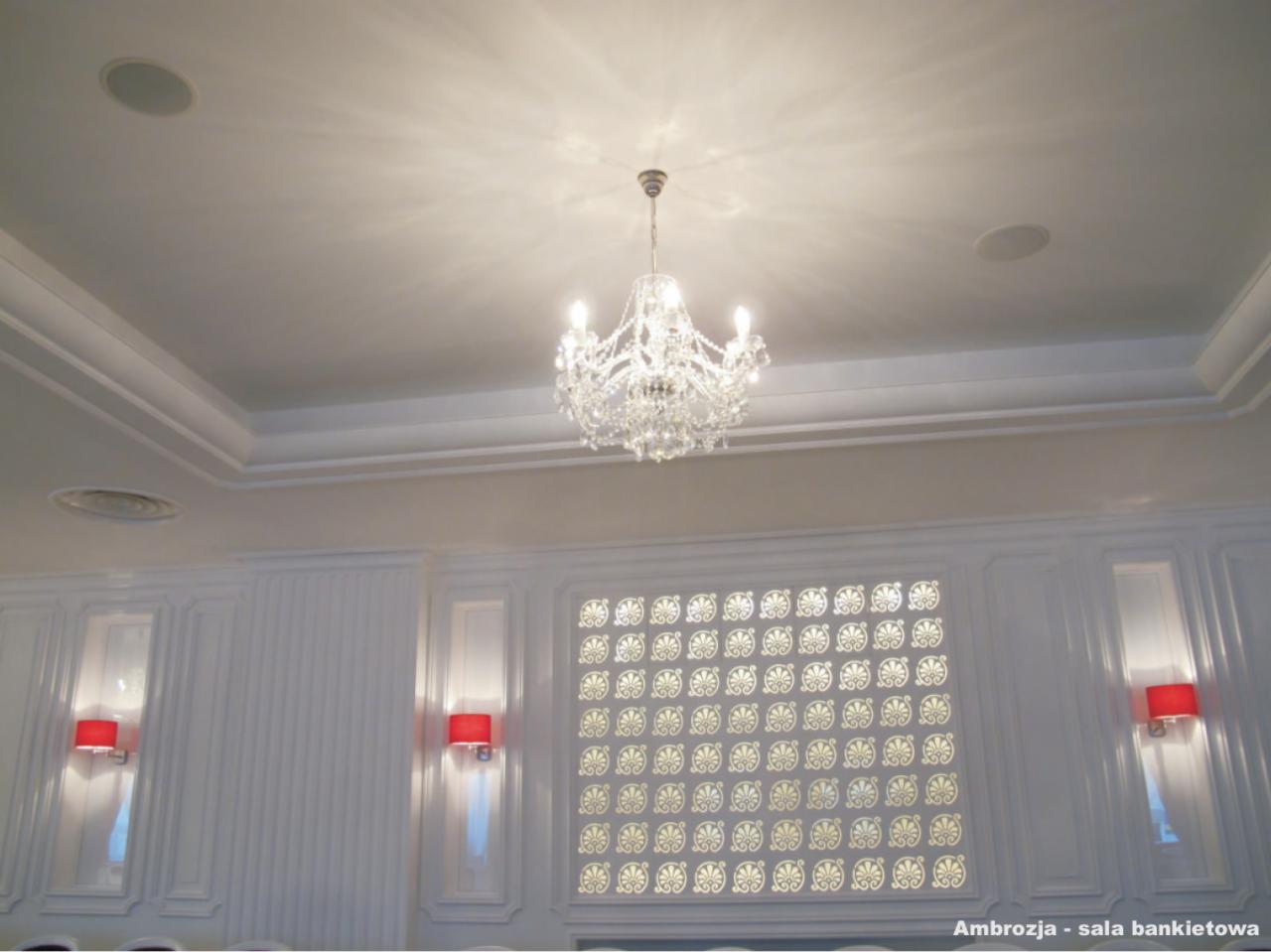 Kryształowy żyrandol oświetla białe wnętrze sali bankietowej z dekoracyjnymi panelami ściennymi i czerwonymi kinkietami.
