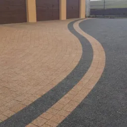 Nowoczesny podjazd z kostki brukowej w odcieniach beżu i szarości, ułożonej w falujący wzór. Widoczne garaże i fragment ogrodzenia w tle.