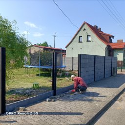 Flis Servis - Montaż betonowego ogrodzenia panelowego: pracownik w czerwonej koszuli układa kostkę brukową przy podstawie słupków, w tle dom z czerwoną dachówką i trampolina.