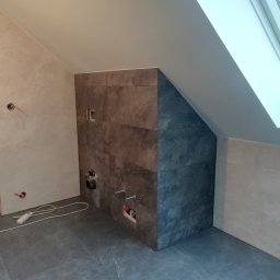 Flis Servis - Łazienka w trakcie remontu: ułożone płytki w odcieniach szarości i brązu na ścianie i podłodze, widoczne otwory na instalacje, przedłużacz na podłodze, okno dachowe w skosie.