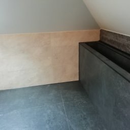 Flis Servis - Wykończona łazienka na poddaszu ze skośnym sufitem, beżowe płytki ścienne imitujące marmur, grafitowa podłoga z płytek i obudowa wanny z ciemnoszarych płytek.