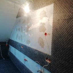 Flis Servis - Ściana łazienki w trakcie prac wykończeniowych: częściowo położona ciemna, trójwymiarowa mozaika, fragmenty ściany z widocznym klejem i przygotowane punkty instalacji elektrycznej i hydraulicznej.