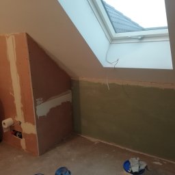 Flis Servis - Poddasze w trakcie wykańczania: ściany częściowo pokryte płytami gipsowo-kartonowymi, widoczne szpachlowanie, okno dachowe z widokiem na dach, rolka papieru toaletowego na ścianie.