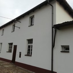 Flis Servis - Odnowiona elewacja budynku z białymi ścianami, brązowym cokołem i orynnowaniem, widoczne okna z białymi ramami i ciemnobrązowe drzwi wejściowe, pochmurne niebo w tle.