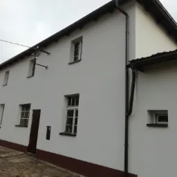 Odnowiona elewacja budynku z białymi ścianami, brązowym cokołem i orynnowaniem, widoczne okna z białymi ramami i ciemnobrązowe drzwi wejściowe, pochmurne niebo w tle.