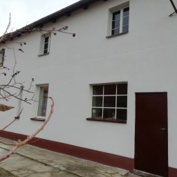 Flis Servis - Biała fasada budynku z brązowymi elementami wykończeniowymi, widoczne okna z białymi ramami, brązowe drzwi wejściowe i fragment drzewa z uschniętymi liśćmi na pierwszym planie.