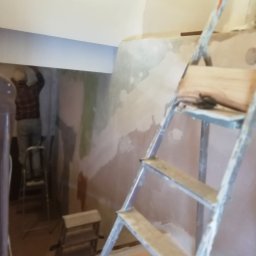 Flis Servis - Pracownik na drabinie szpachluje nierówną ścianę w pomieszczeniu w trakcie remontu, widoczna druga drabina i narzędzia