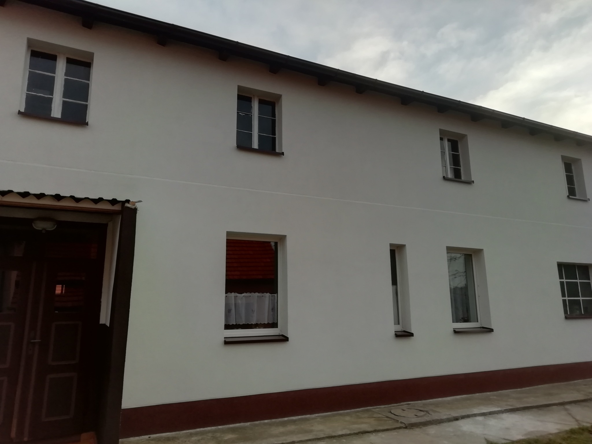 Dwukondygnacyjny budynek z białymi ścianami i kilkoma oknami o różnej wielkości, widoczny z zewnątrz w pochmurny dzień.