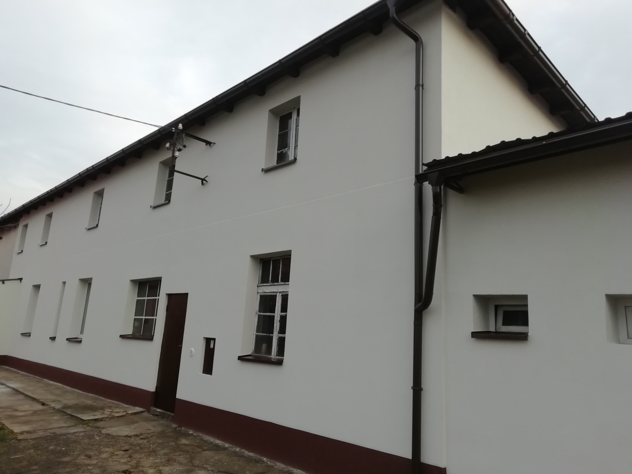 Odnowiona elewacja budynku z białymi ścianami, brązowym cokołem i orynnowaniem, widoczne okna z białymi ramami i ciemnobrązowe drzwi wejściowe, pochmurne niebo w tle.