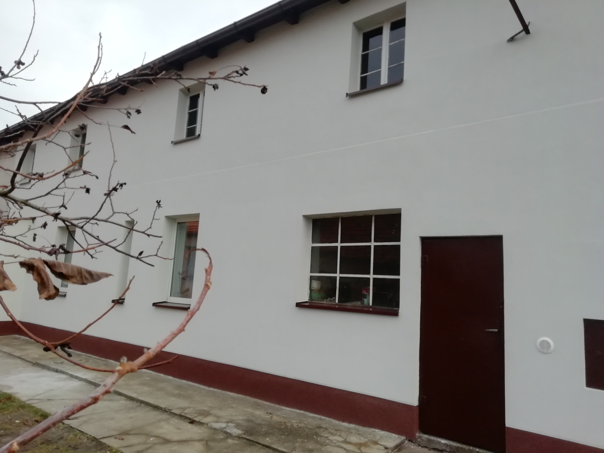 Biała fasada budynku z brązowymi elementami wykończeniowymi, widoczne okna z białymi ramami, brązowe drzwi wejściowe i fragment drzewa z uschniętymi liśćmi na pierwszym planie.