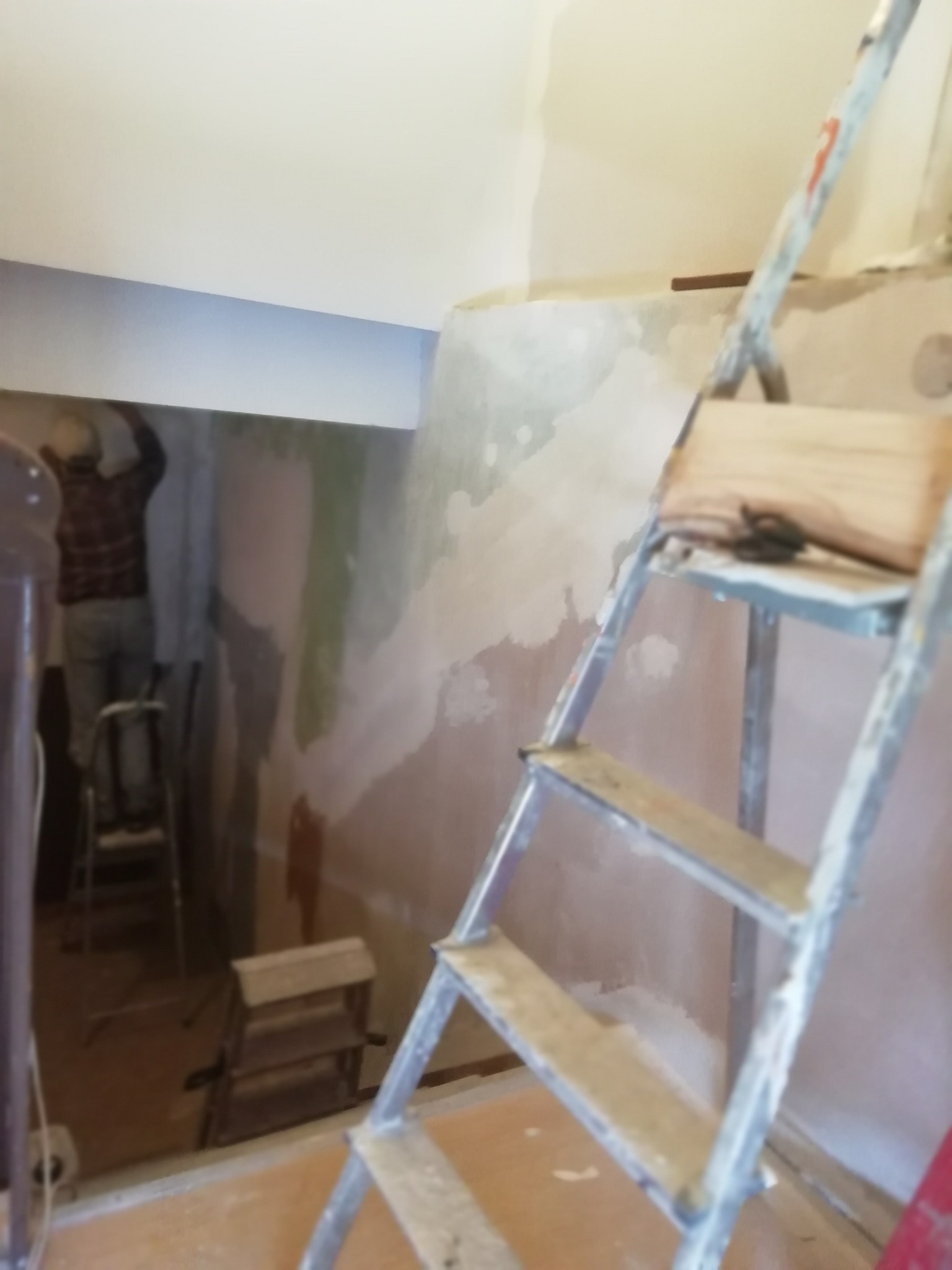 Pracownik na drabinie szpachluje nierówną ścianę w pomieszczeniu w trakcie remontu, widoczna druga drabina i narzędzia