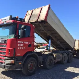 Czerwona ciężarówka Scania z podniesioną skrzynią ładunkową, widoczny hydrauliczny siłownik pod skrzynią, na kabinie logo DSV Transport, w tle inne pojazdy i błękitne niebo.