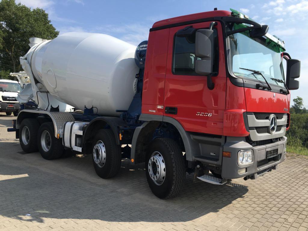 Czerwona betonomieszarka Mercedes-Benz 3236 na kostce brukowej, z widocznym bębnem mieszającym i kabiną kierowcy, zaparkowana w słoneczny dzień.
