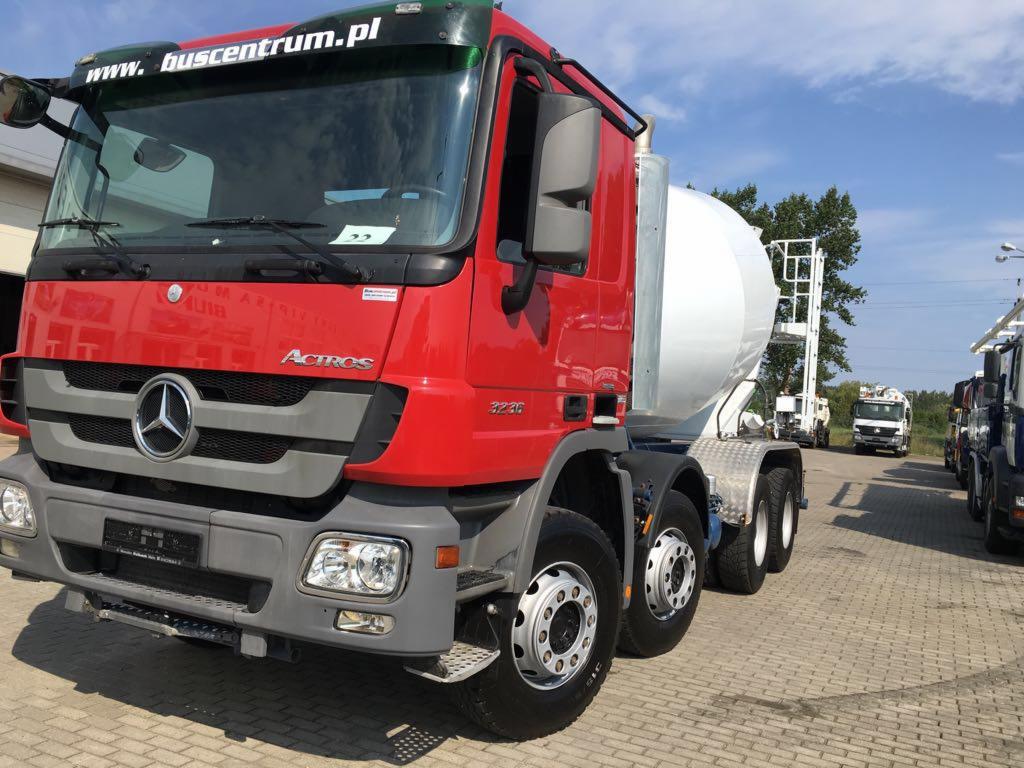 Czerwona betonomieszarka Mercedes Actros 3236 z białą gruszką, zaparkowana na placu z kostki brukowej, widok z przodu, w tle inne pojazdy.