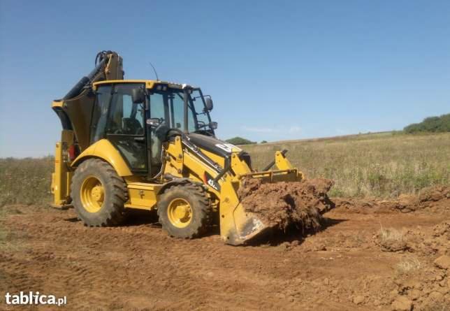 Żółta koparko-ładowarka Caterpillar 434E w trakcie prac ziemnych na polu, z łyżką wypełnioną ziemią; widoczne ślady opon i naruszona gleba.