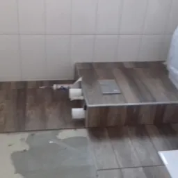 Montaż toalety na podwyższeniu z płytek imitujących drewno, widoczne akcesoria montażowe i białe płytki podłogowe w trakcie układania.