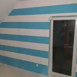 Ściana pokoju na poddaszu pomalowana w poziome, biało-błękitne pasy, z widocznym oknem w białej ramie i gniazdkami elektrycznymi przy podłodze.