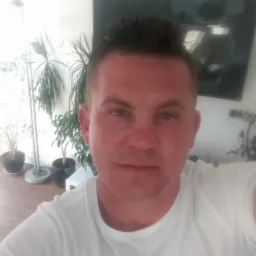 Mężczyzna w białej koszulce na tle jasnego wnętrza z roślinami i telewizorem. Zdjęcie z bliska, oświetlenie naturalne.