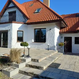 Biały dom z czerwoną dachówką i balkonem. Teksturowana elewacja, kamienne schody, lampy ogrodowe i dekoracyjne rośliny tworzą spójny, estetyczny wygląd.