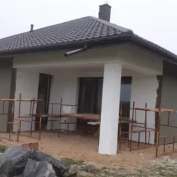 Parterowy dom jednorodzinny z ciemną dachówką, częściowo otynkowany, z widocznymi rusztowaniami i workami na ziemi, w trakcie prac elewacyjnych.