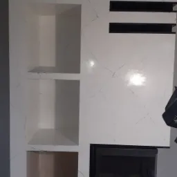 Biała ściana z wbudowanym kominkiem i półkami, wykończona w stylu imitującym marmur z czarnymi żyłkami, nowoczesny design.