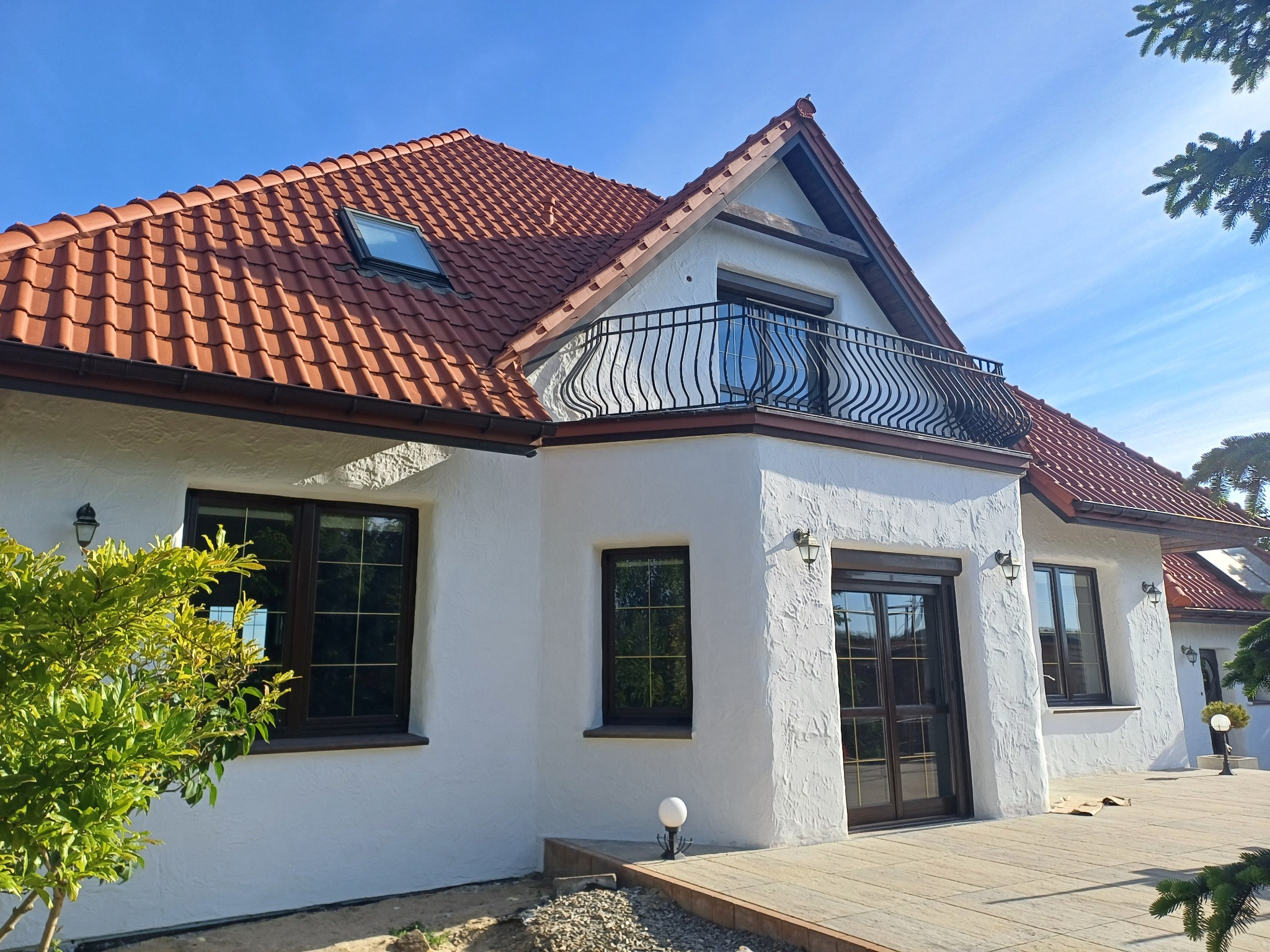 Elegancki dom z balkonem i czerwoną dachówką w słoneczny dzień. Biała fasada z oknami w ciemnych ramach. Widoczny fragment tarasu.