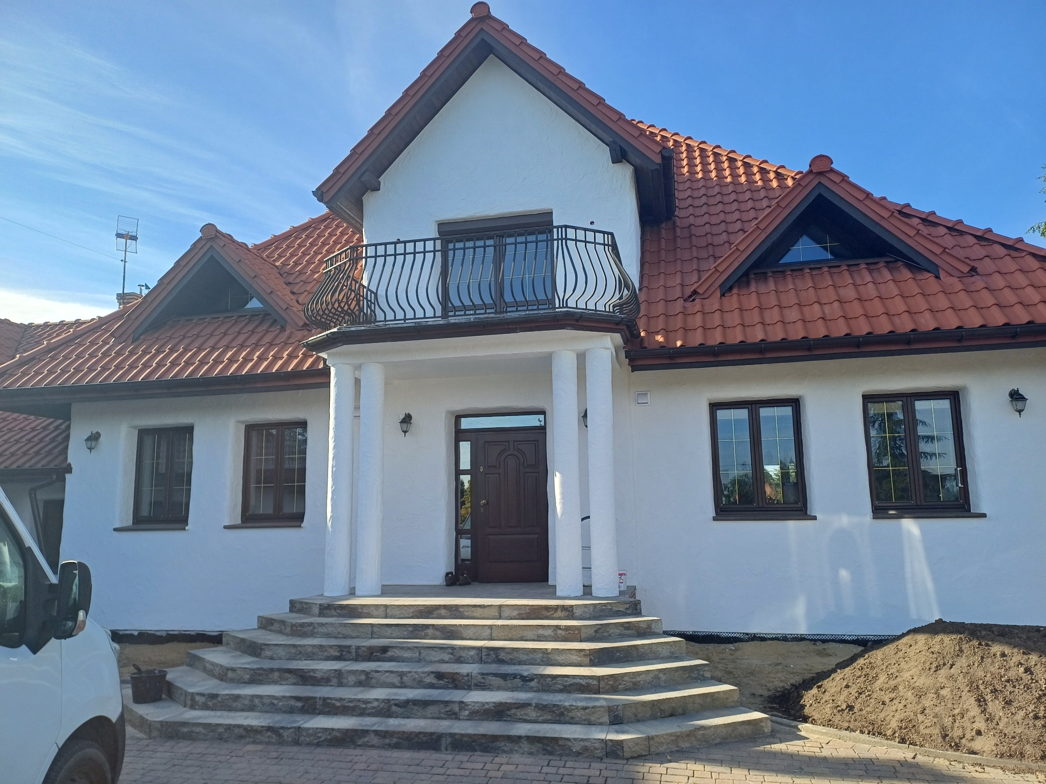 Biały dom z czerwoną dachówką i balkonem z czarną balustradą, widok z dołu. Uwagę przyciągają schody wejściowe z kostki brukowej i faktura elewacji. Częściowo widoczny samochód.