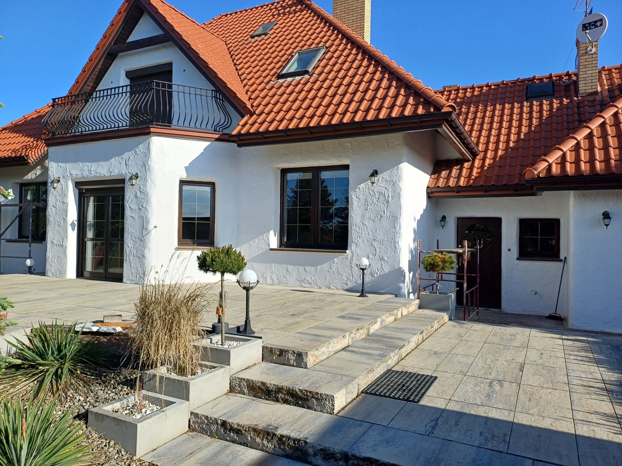 Biały dom z czerwoną dachówką i balkonem. Teksturowana elewacja, kamienne schody, lampy ogrodowe i dekoracyjne rośliny tworzą spójny, estetyczny wygląd.