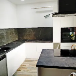 Nowoczesna, jasna kuchnia z białymi szafkami, czarnym blatem z efektem kamienia i pasującym panelem ściennym. Na blacie czajnik z drewnianą rączką, a w tle piekarnik z włączonym oświetleniem.