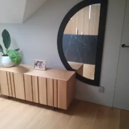 Drewniana komoda z ryflowanymi frontami, na której stoi roślina doniczkowa i ramka ze zdjęciem, obok wisi półokrągłe lustro w czarnej ramie, odbijające fragment blatu z czarnego marmuru.