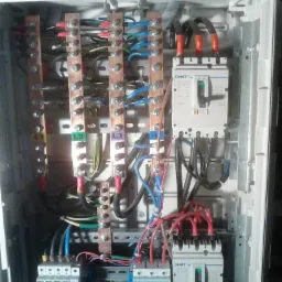 Otwarta skrzynka rozdzielcza z widocznymi bezpiecznikami, szynami zbiorczymi oznaczonymi L1, L2, L3 i przewodami w różnych kolorach, zamontowana wewnątrz białej obudowy.