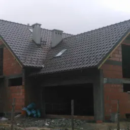 Murowany dom w trakcie budowy z widoczną czerwoną cegłą, szarą dachówką i elementami betonowymi. Widoczne otwory okienne i drzwiowe. Surowy stan budynku.