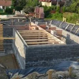 Zalane fundamenty domu w trakcie budowy, z widoczną konstrukcją szalunkową i zbrojeniem, na tle zieleni i zabudowań. Etap prac fundamentowych.