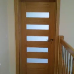 Dębowa, fornirowana ościeżnica z przesuwnymi, matowymi panelami. Widoczny fragment balustrady z drewna w ciepłym odcieniu. Minimalistyczny design.